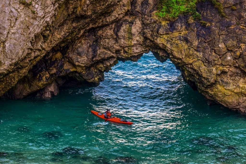 Kayaker in a red sea kayak beneath a coastal rock arch