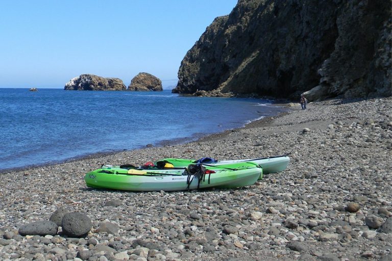Two green sit-on-top kayaks on a pebble beach below cliffs