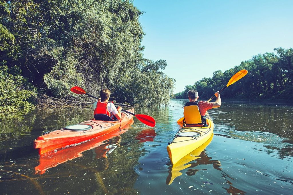 Two kayakers paddling red and yellow kayaks along a tree-lined river